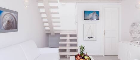 A modern, white-themed living room with a white sofa, a coffee table with fruit, a staircase, and a door with a sailboat painting.