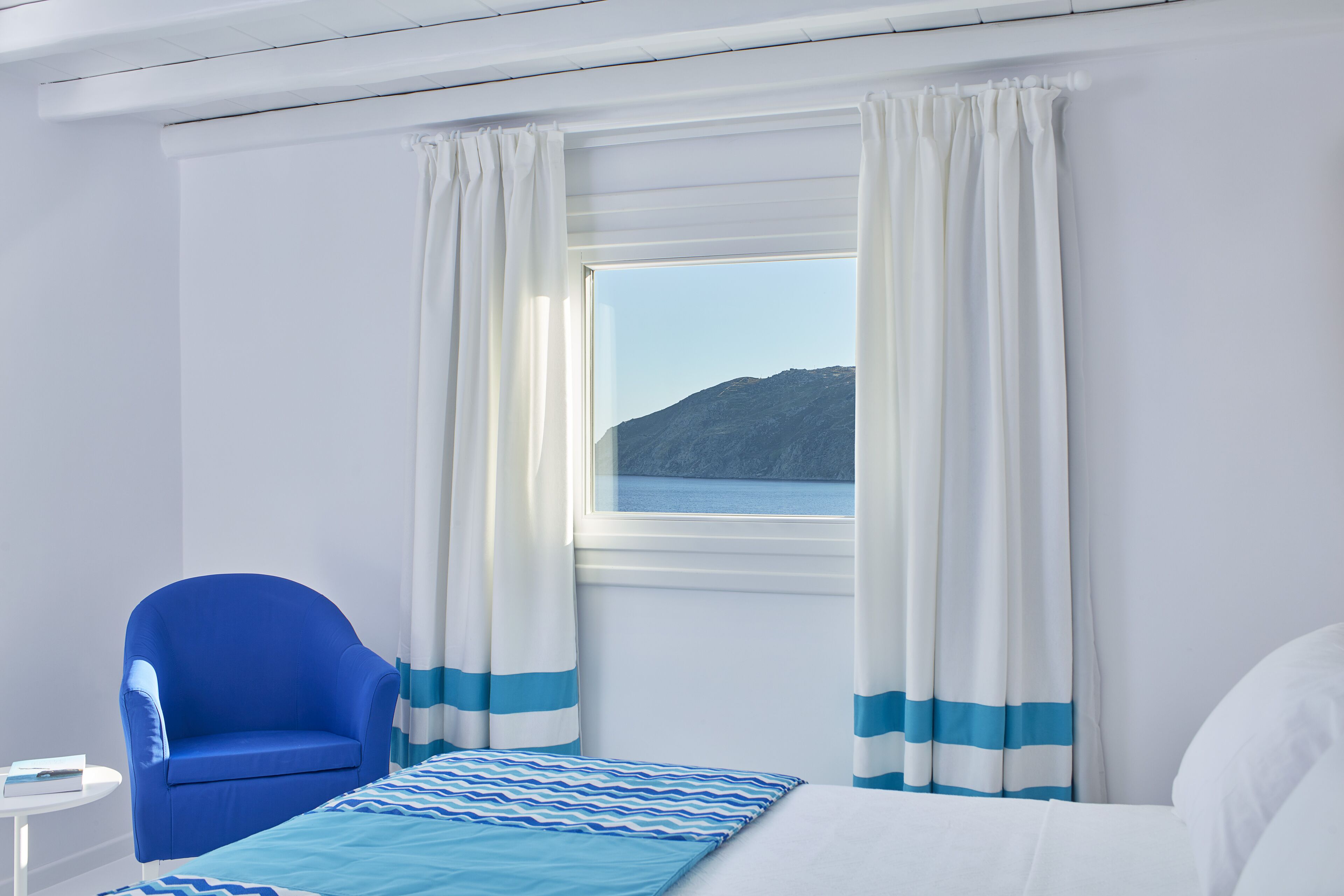 Una habitación con una cama, una silla azul, una mesita y una ventana con cortinas.