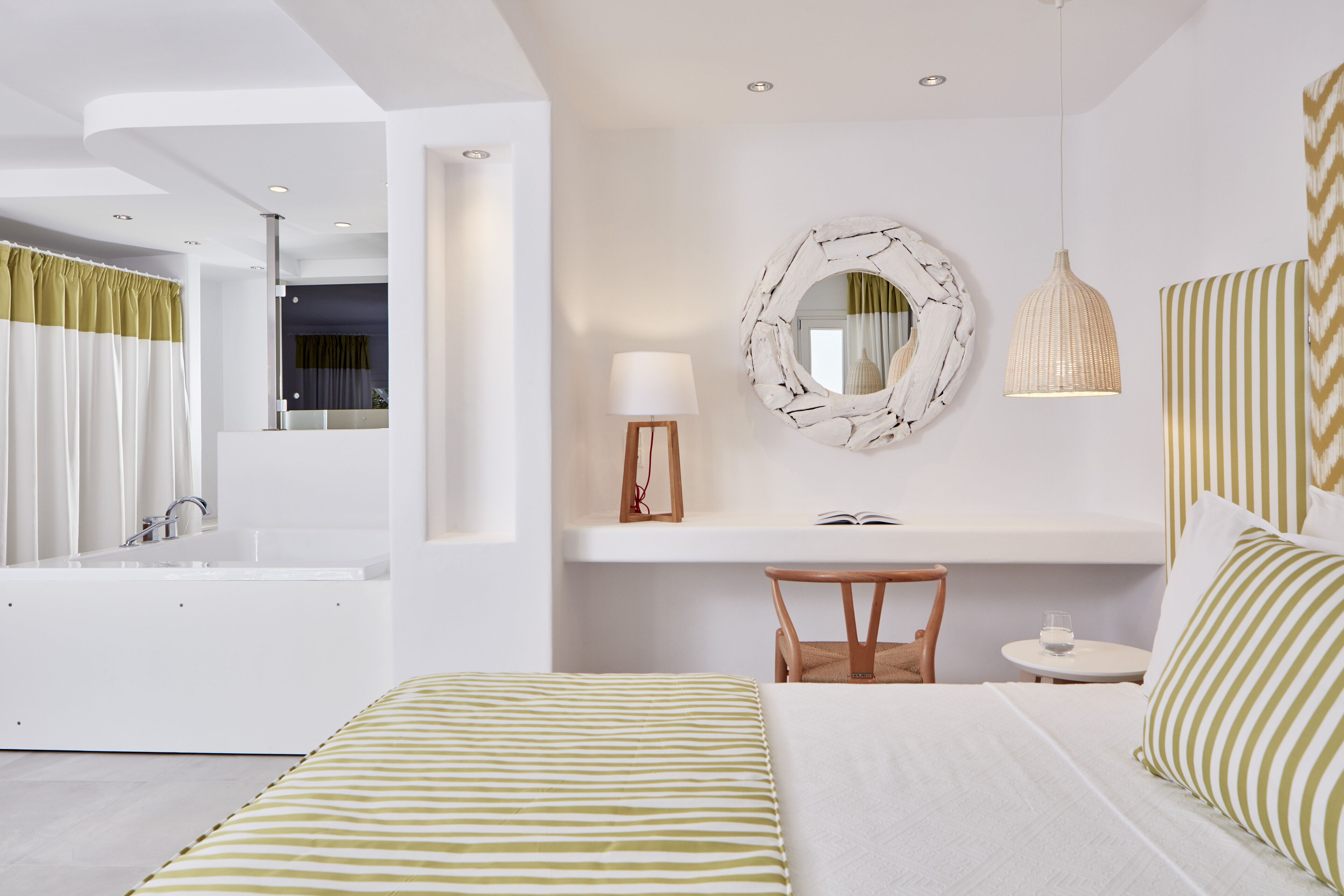 A modern hotel room with a large bed, a sitting area with a small table and chair, a vanity with a mirror, and a bathtub.