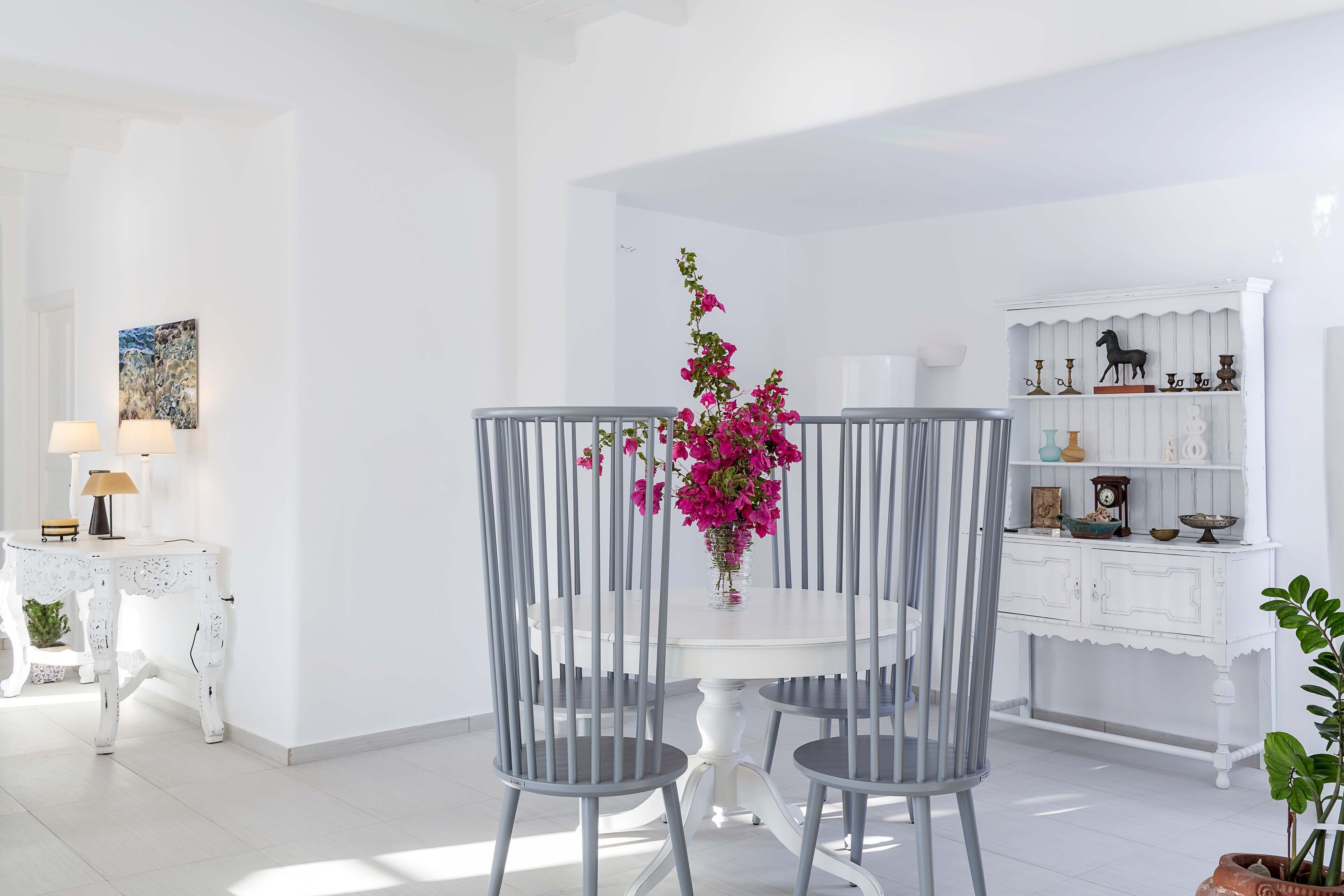 A dining area with a white table and grey chairs, a vase with pink flowers, and a white cabinet with decorative items.