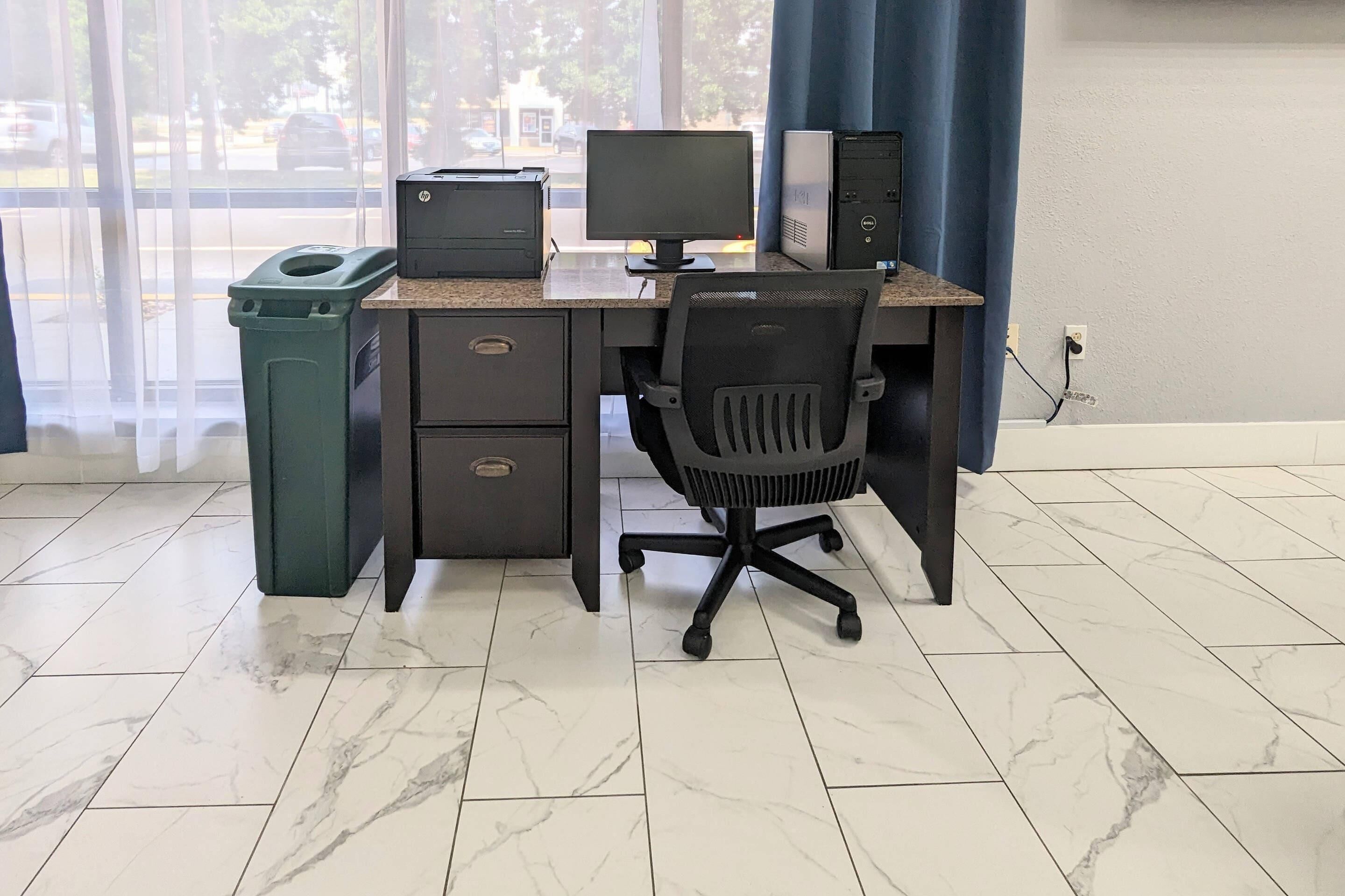 Un espacio de trabajo con una computadora de escritorio, un monitor e impresora sobre un escritorio, un cubo de basura y una silla.