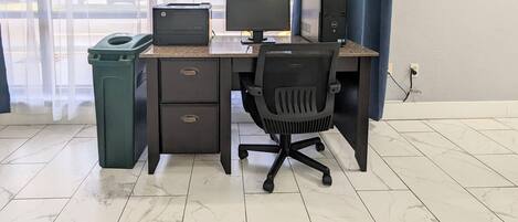 Un espacio de trabajo con una computadora de escritorio, un monitor e impresora sobre un escritorio, un cubo de basura y una silla.