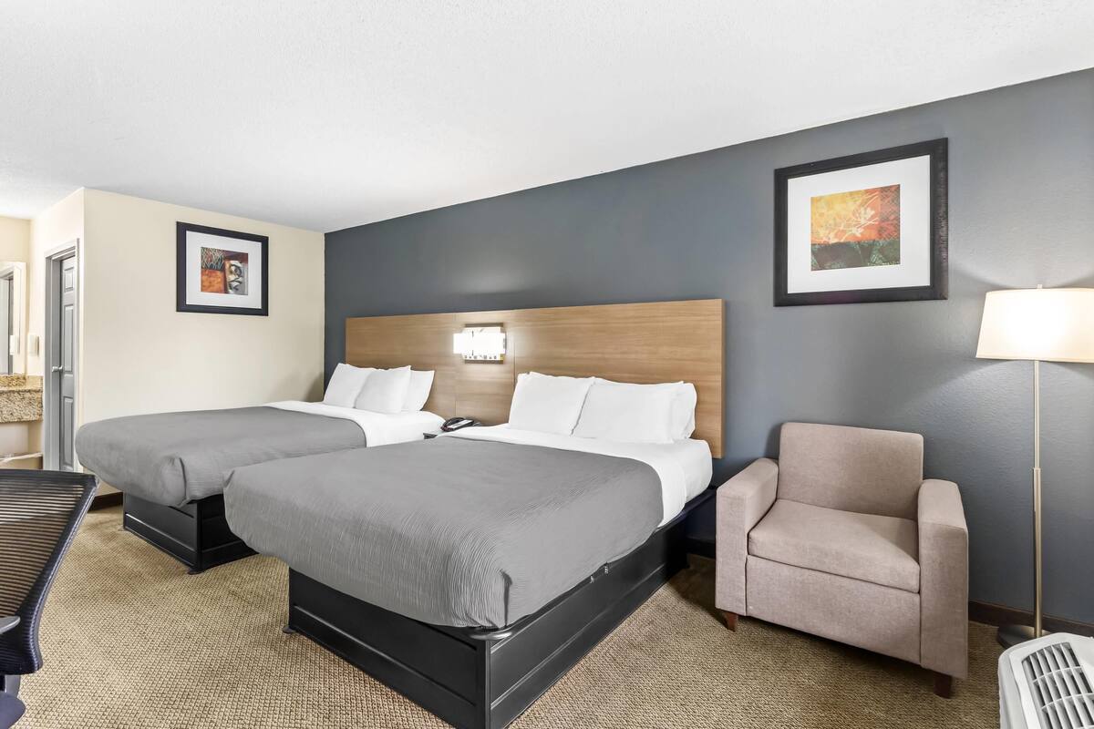 A hotel room with two beds, a chair, a desk, and a lamp.