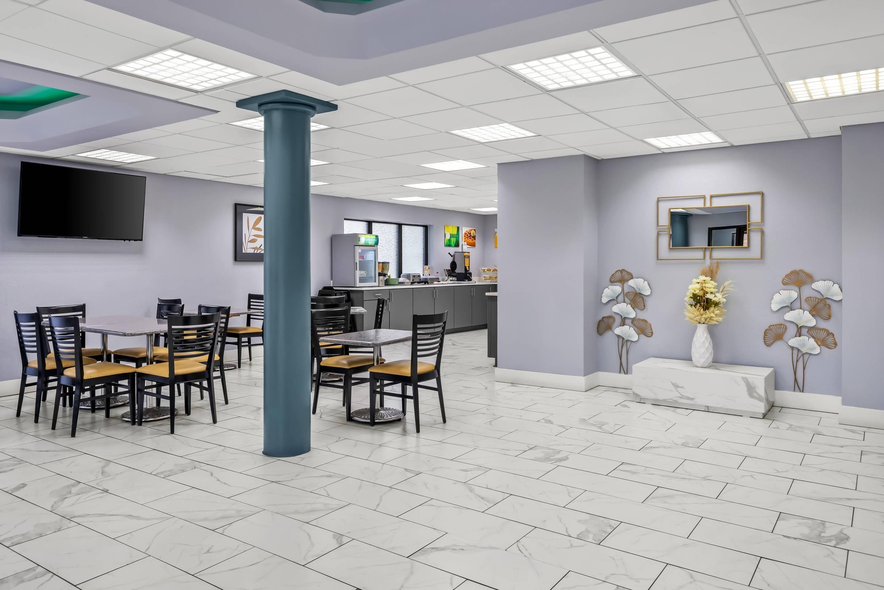 A hotel dining area with marble flooring, a central pillar, a flat-screen TV, and a reception desk in the background.