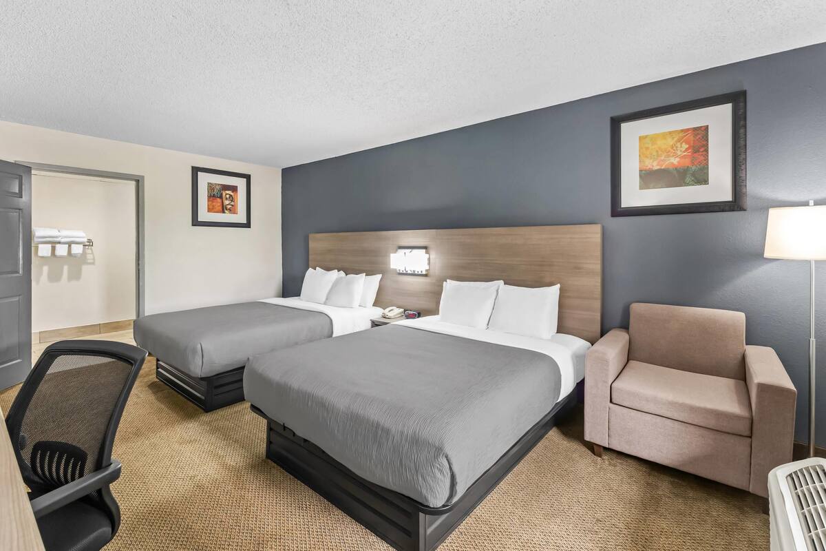 A hotel room with two beds, a desk, a chair, and a lamp.