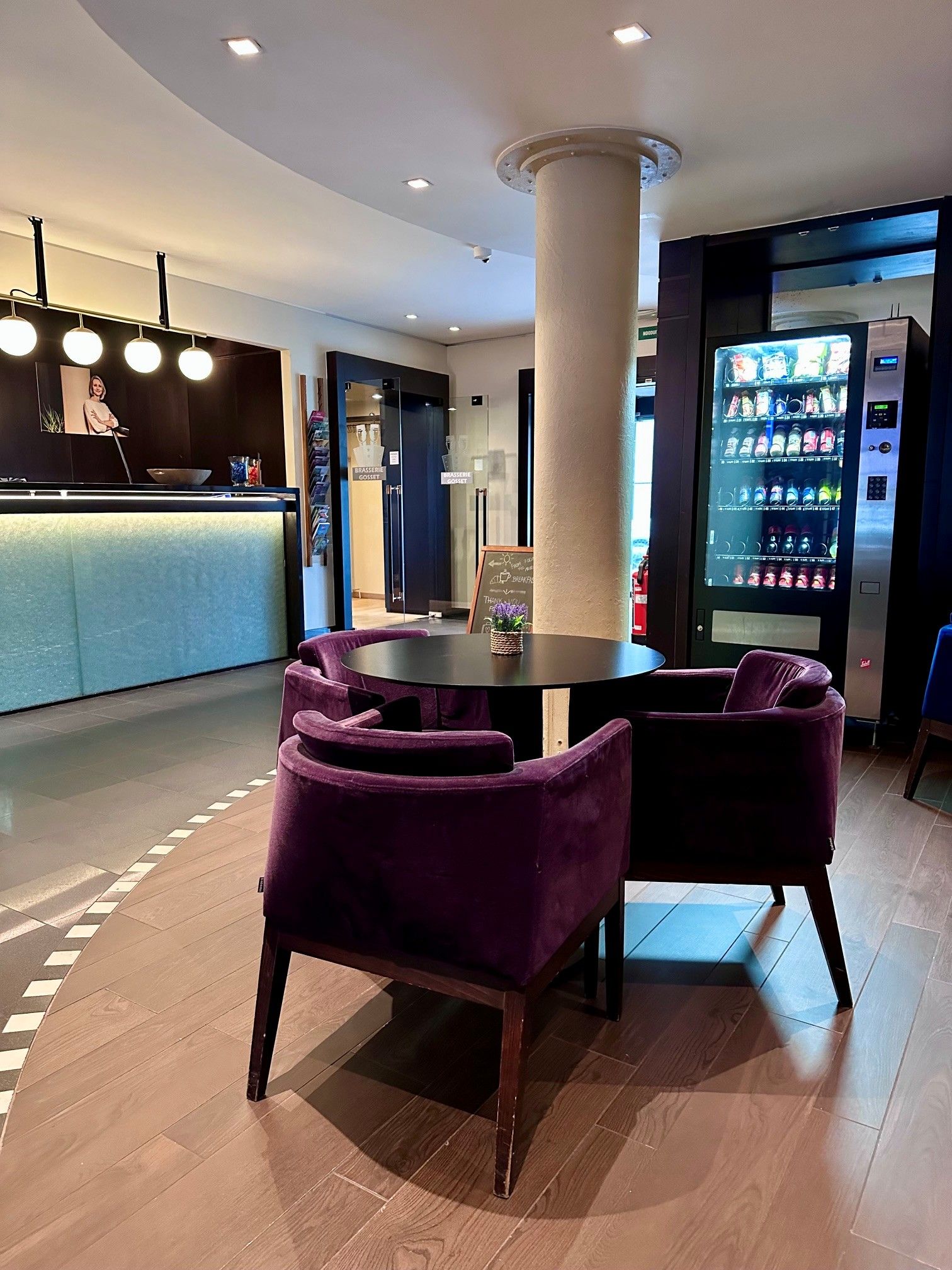 Ein modernes Interieur mit einem runden Tisch und violetten Stühlen, einem Verkaufsautomaten und einem Empfangsbereich mit Theke und Spiegel.
