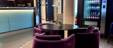 Un interior moderno con una mesa redonda y sillas moradas, una máquina expendedora y una zona de recepción con mostrador y espejo.