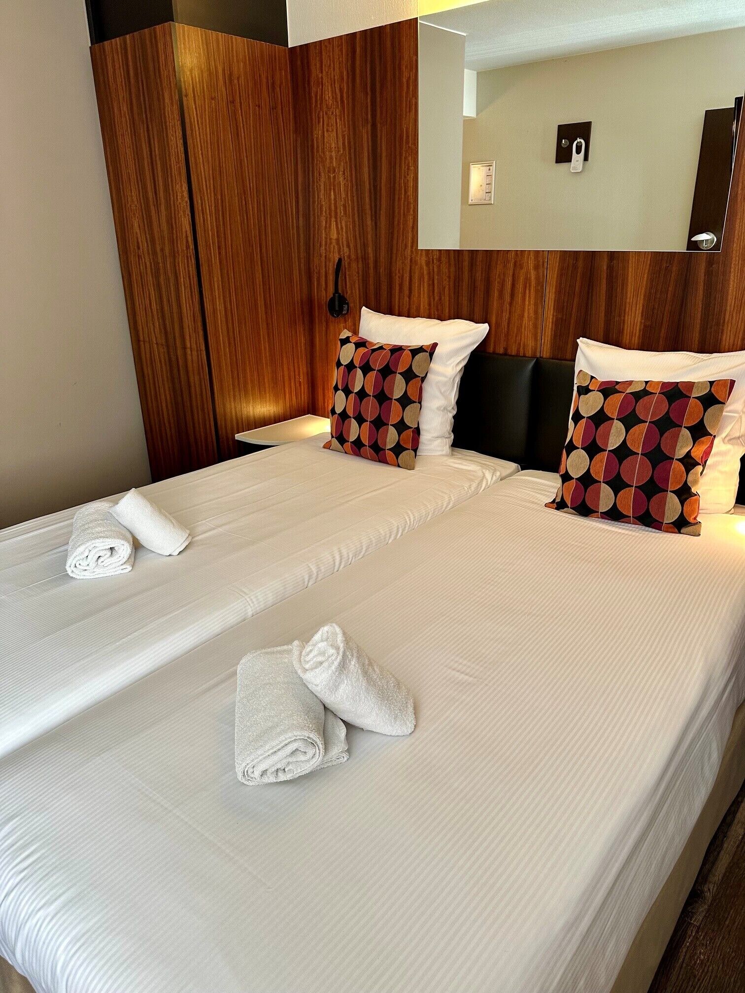 A double bed with white linens and two decorative pillows, a wooden headboard, and a bedside table with a lamp.