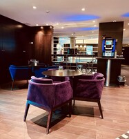 Un moderno vestíbulo de hotel con una mesa redonda y sillas moradas, una zona de bar con una máquina de café, y vistas de la ciudad a través de amplios ventanales.