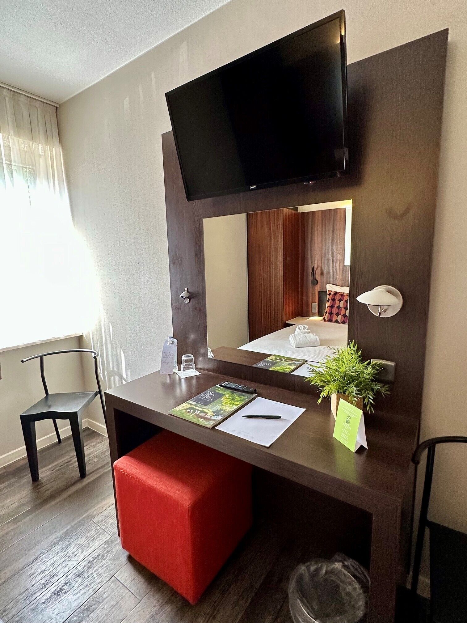 Habitación de hotel con una televisión de pantalla plana montada sobre un escritorio de madera, un reposapiés rojo, una pequeña planta en maceta y un espejo que refleja una cama y cortinas.