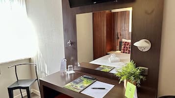 Ein Hotelzimmer mit Flachbildfernseher an der Wand über einem Holztisch, einem roten Hocker, einer kleinen Topfpflanze und einem Spiegel, der ein Bett und Vorhänge reflektiert.