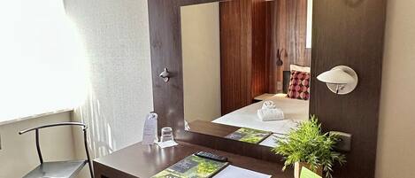 Habitación de hotel con una televisión de pantalla plana montada sobre un escritorio de madera, un reposapiés rojo, una pequeña planta en maceta y un espejo que refleja una cama y cortinas.