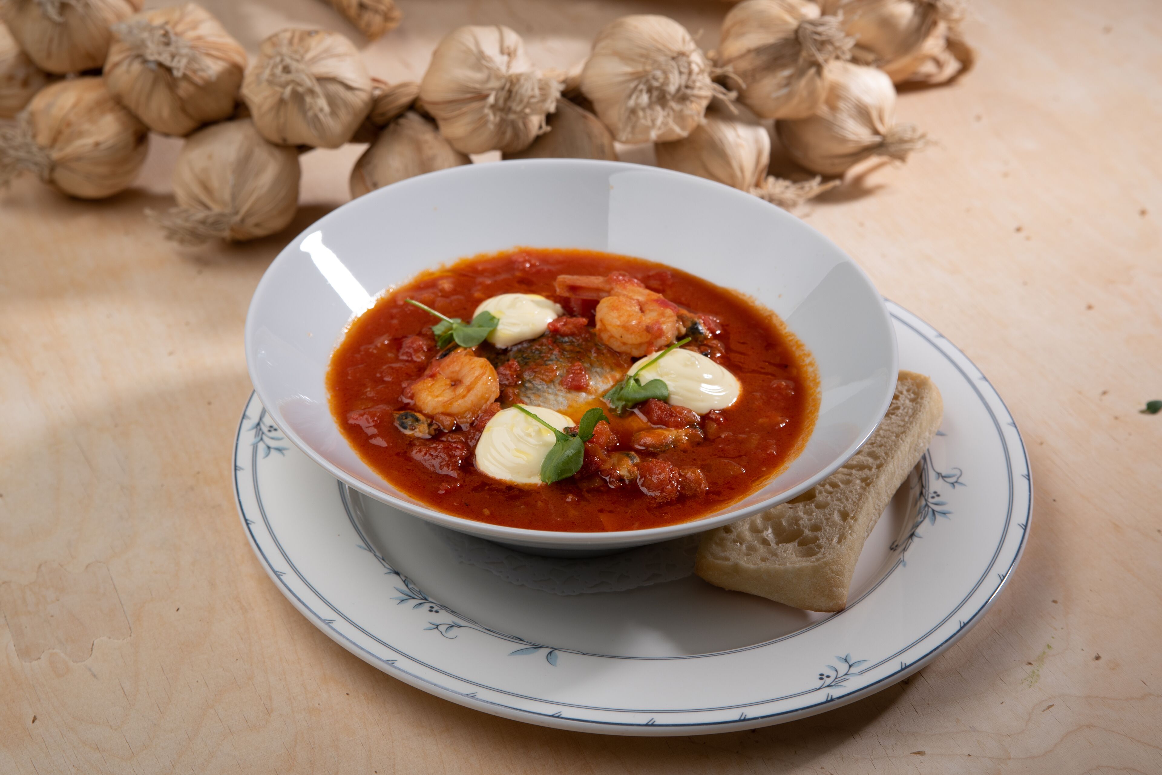 Un tazón de sopa con pan sobre un plato, rodeado de bulbos de ajo.