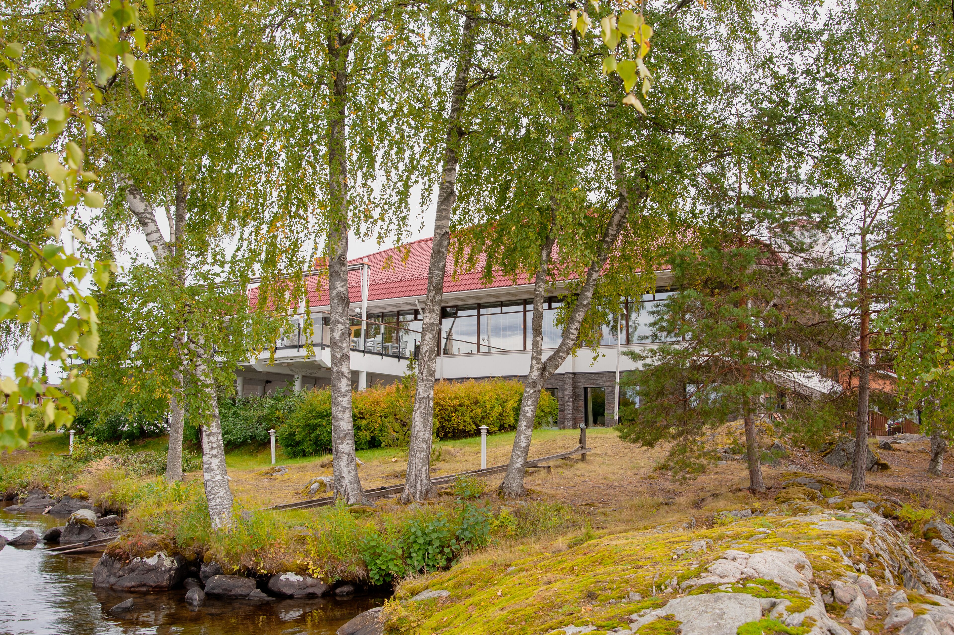 A modern house with a red roof, surrounded by trees and a small stream.