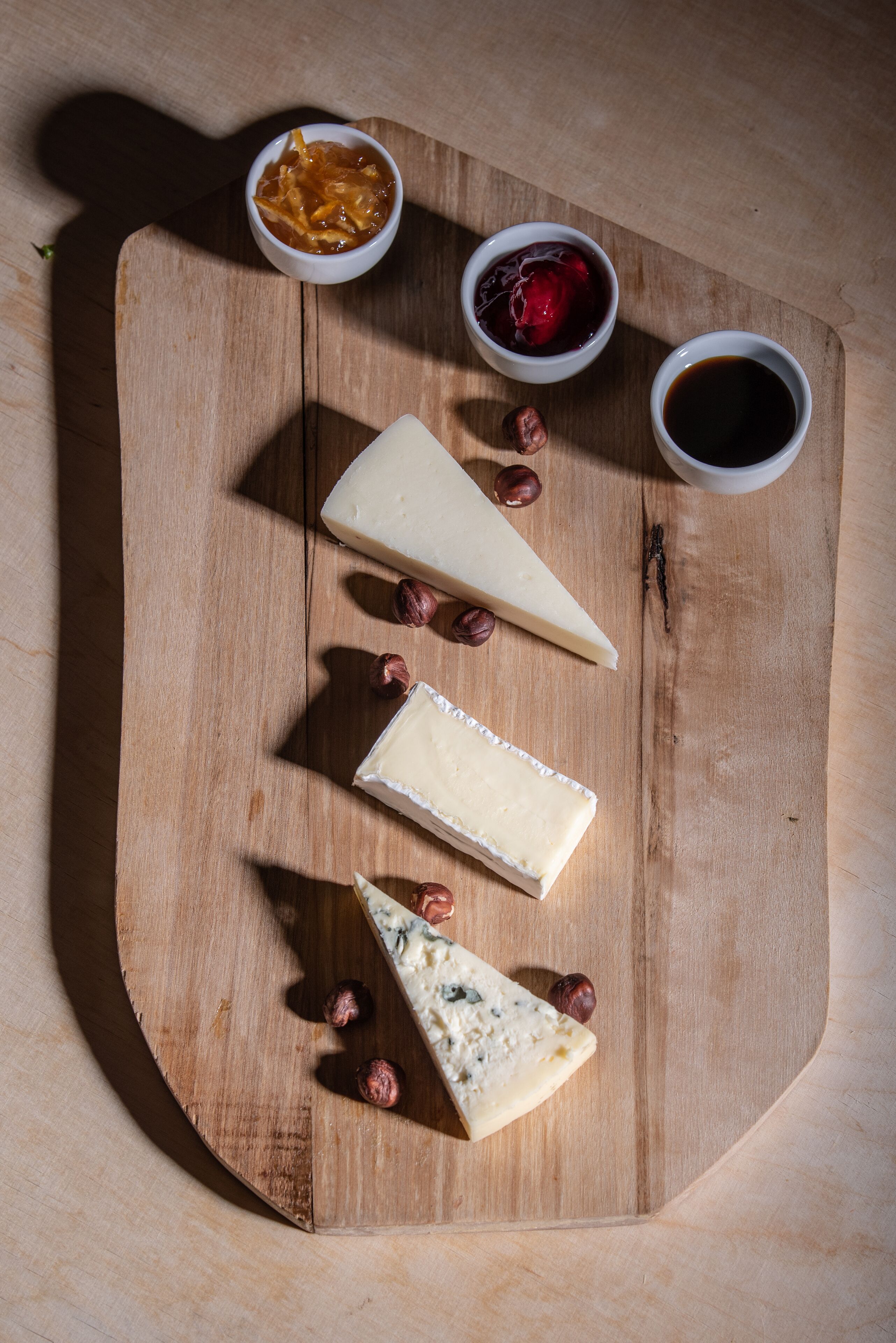 A wooden board with three types of cheese, two small bowls of condiments, and scattered nuts.