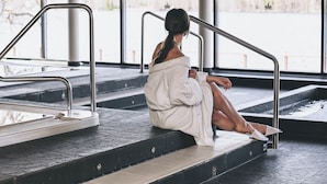 A person sitting on a spa bench by a poolside, with a view of a calm water body and a building in the background. - Kumpeli Spa (Heinola)