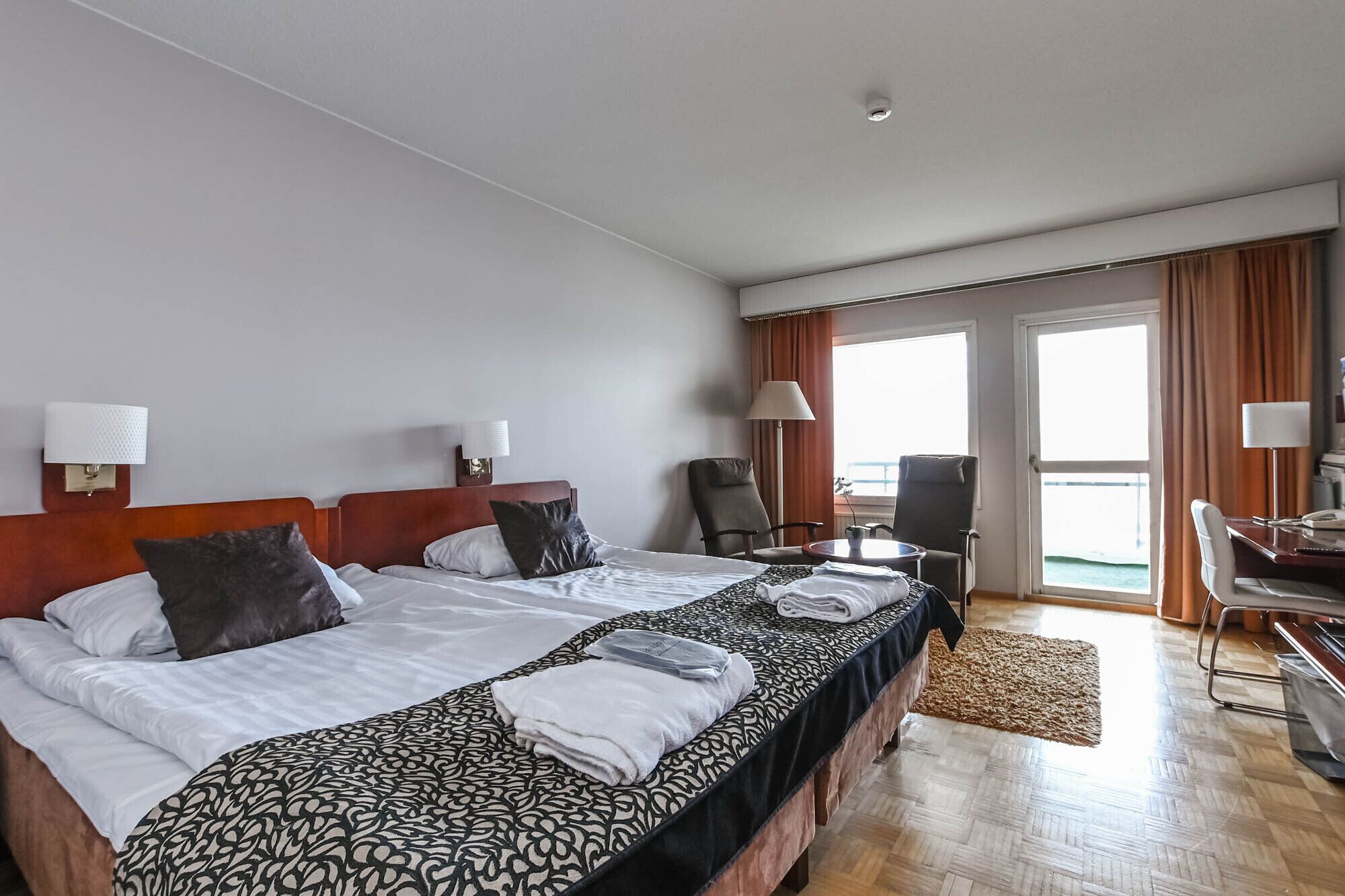 A hotel room with a large bed, two bedside tables, a desk, a chair, and a view of the sea through the windows.