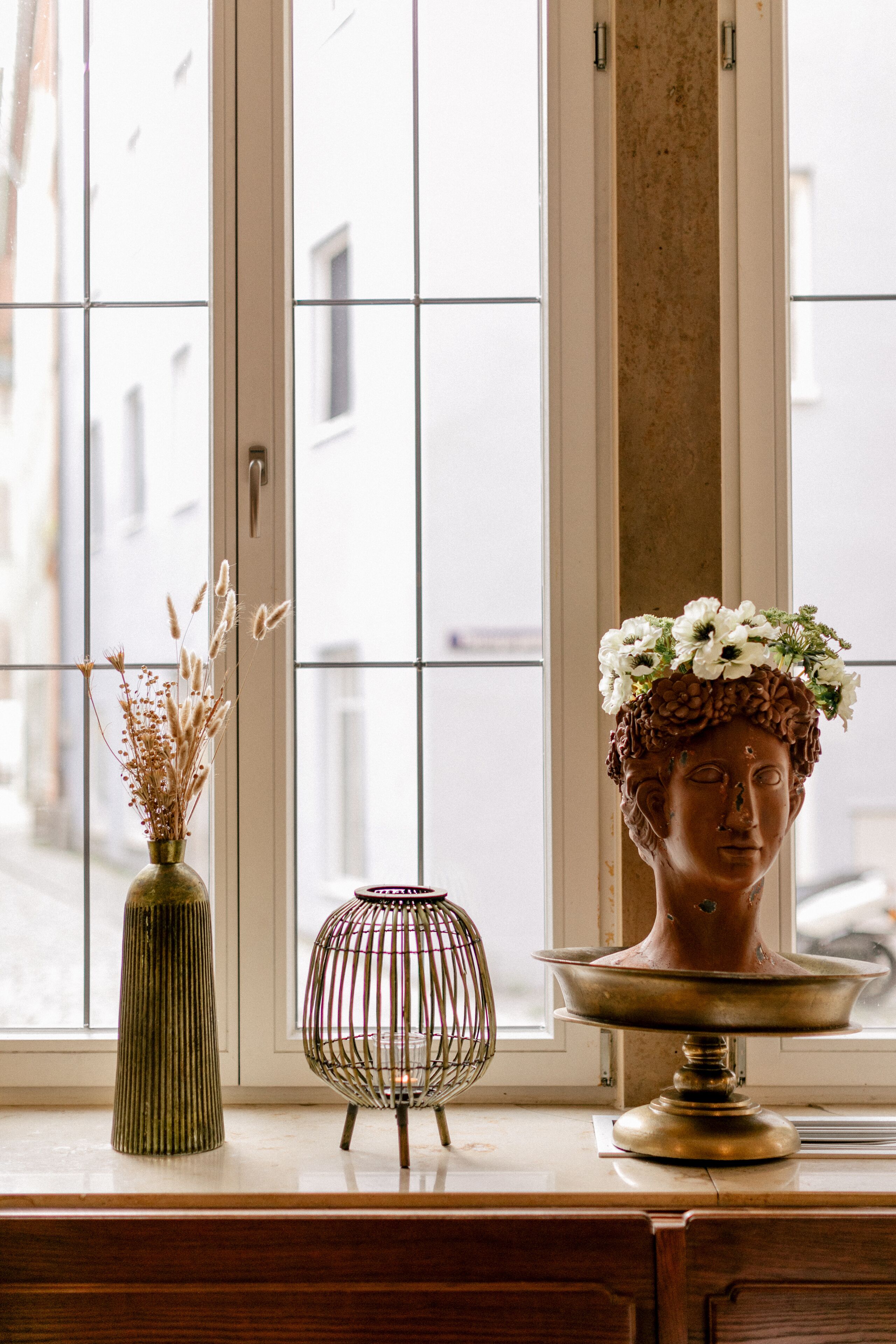 Une fenêtre donnant sur des immeubles, un vase contenant des fleurs séchées et un buste posé sur un piédestal.