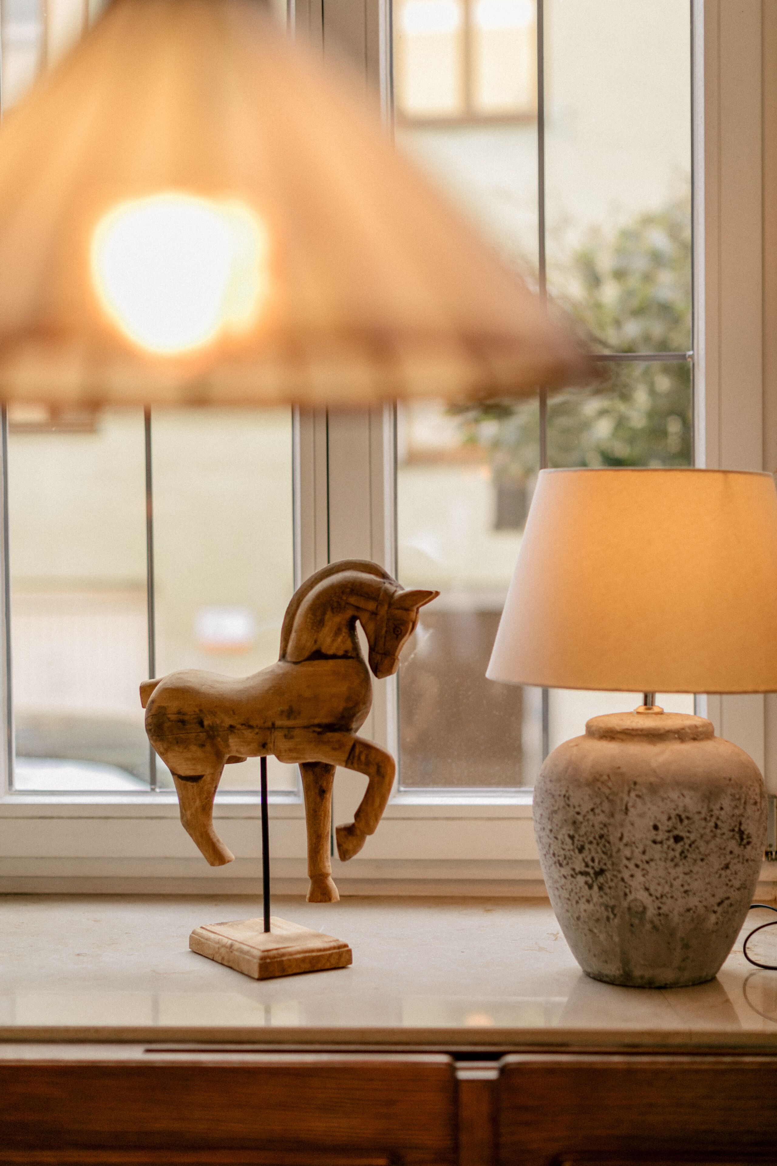 Een houten paardbeeldje op een voetstuk naast een lamp op een raamdorpel.
