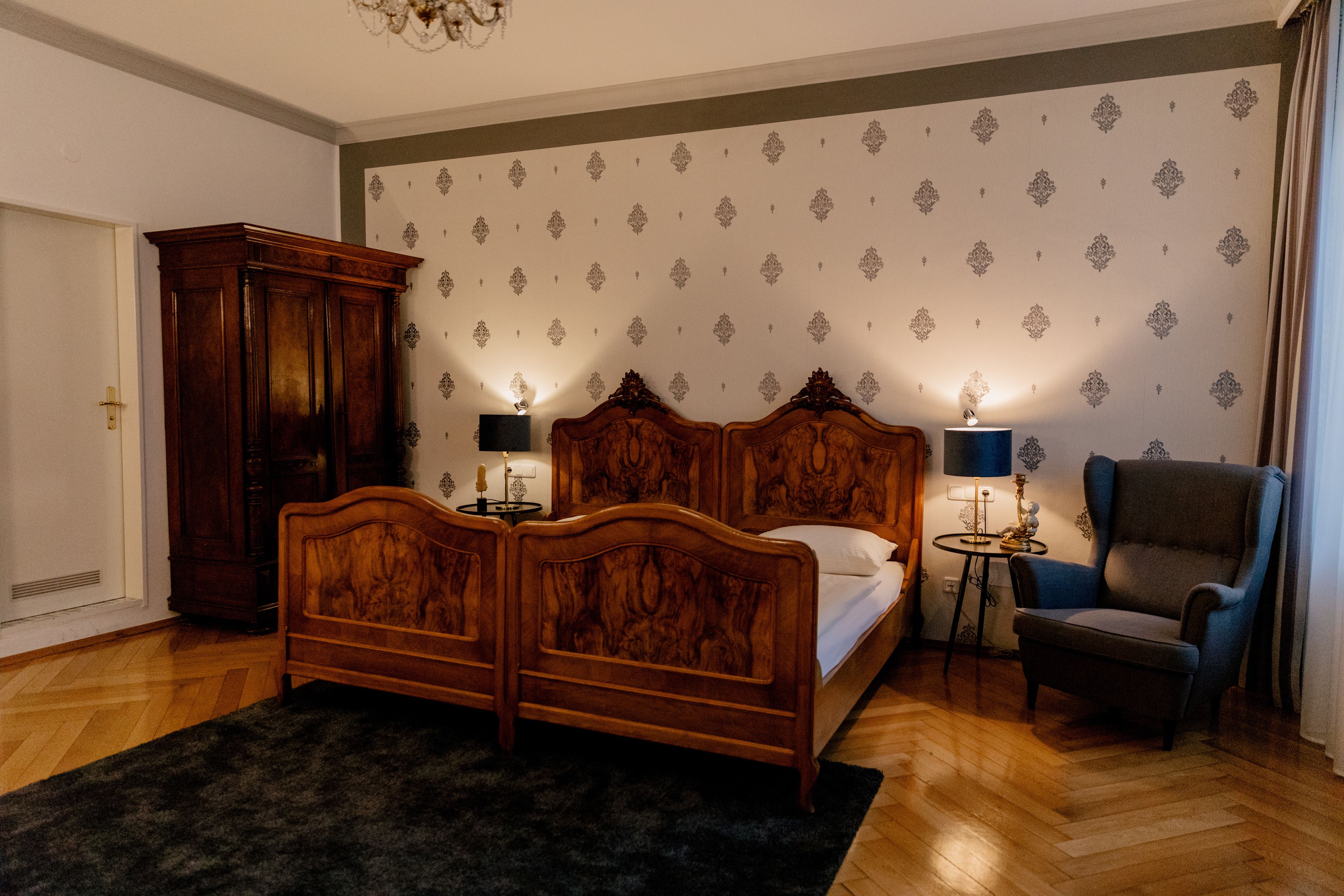 Quarto com cama de madeira, duas mesas de cabeceira com luminárias, uma poltrona azul e um tapete escuro.