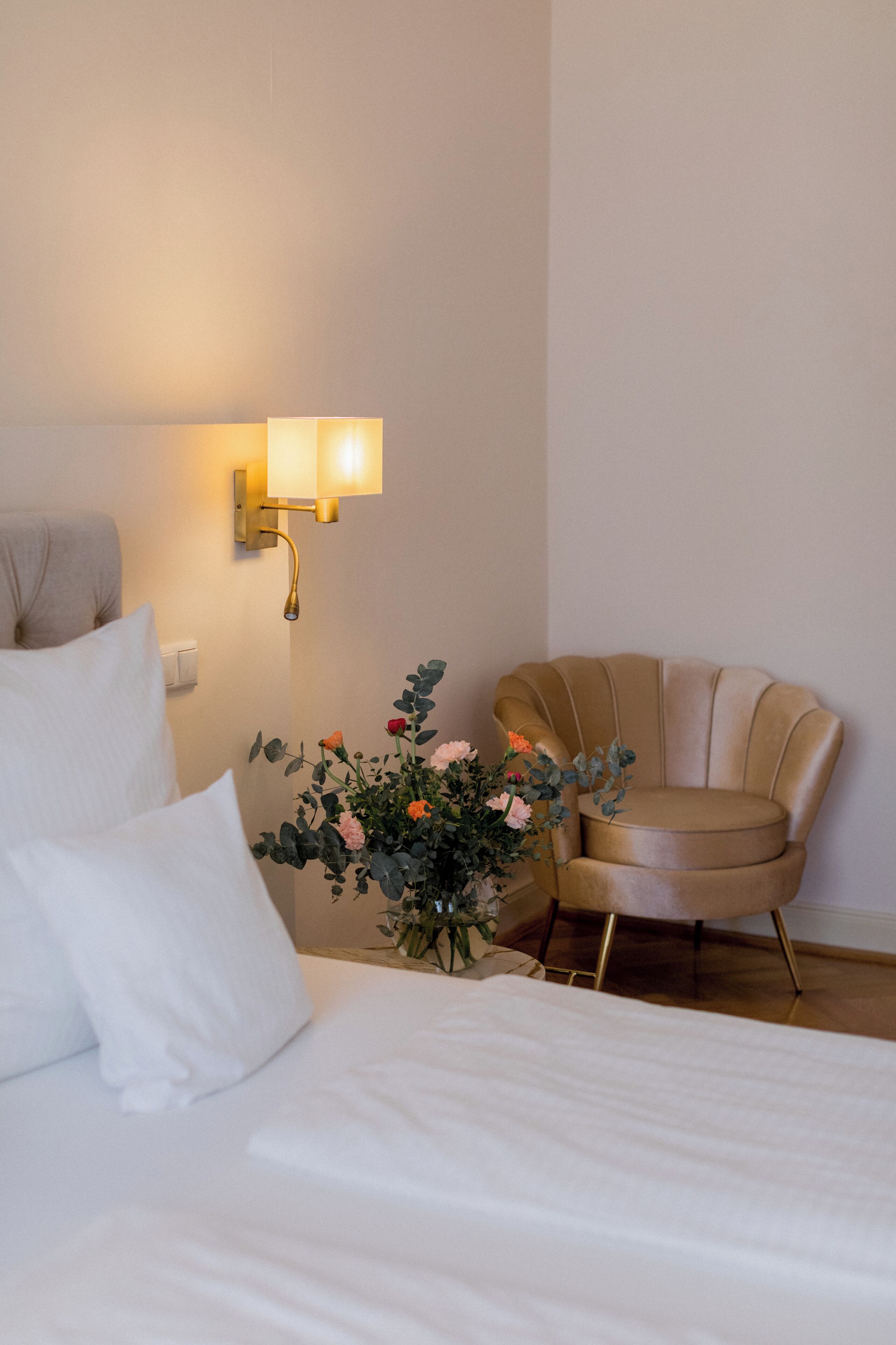 Una habitación de hotel con una cama, una silla, una lámpara de pared y un florero con flores.