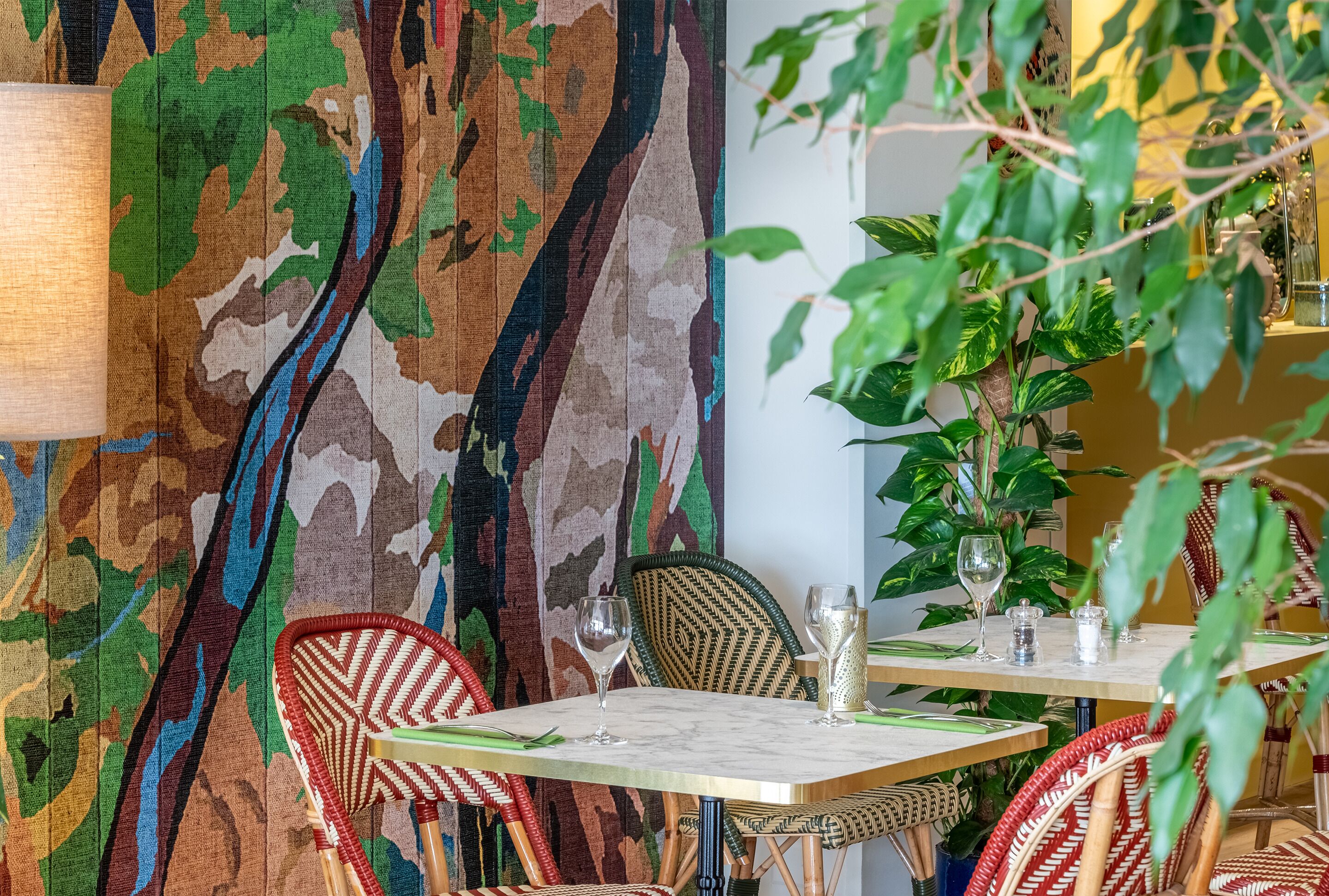 A dining area with a patterned wall, wicker chairs, and a table set for two with glasses and cutlery.