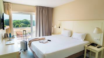 Een hotelkamer met een bed, een bureau met een laptop, een koffer, een televisie en een balkon met gordijnen.