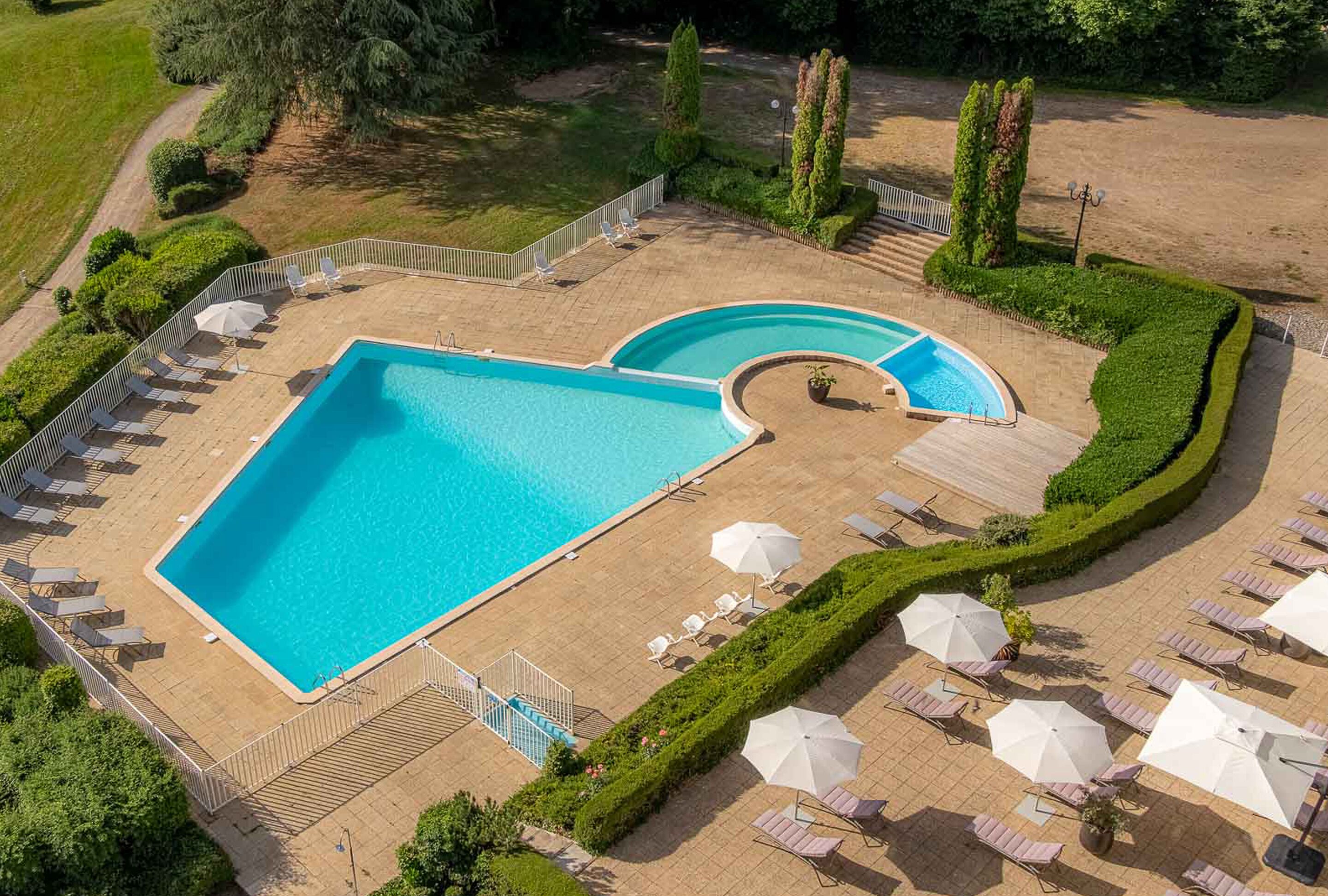 An aerial view of a swimming pool area with two pools, surrounded by a well-maintained garden and a paved deck with lounge chairs and umbrellas.