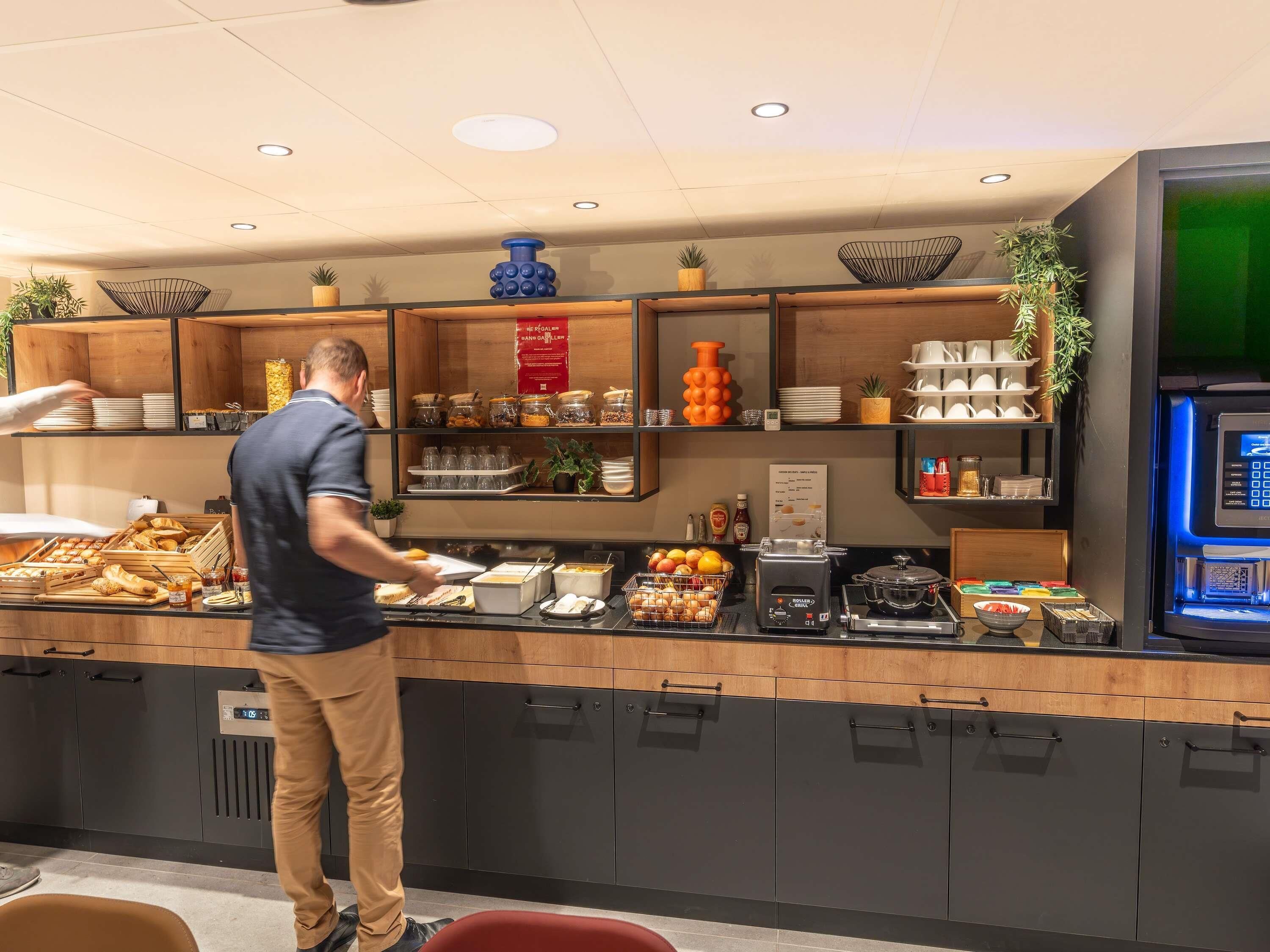A breakfast buffet area with various food items, a coffee machine, and a digital menu board.
