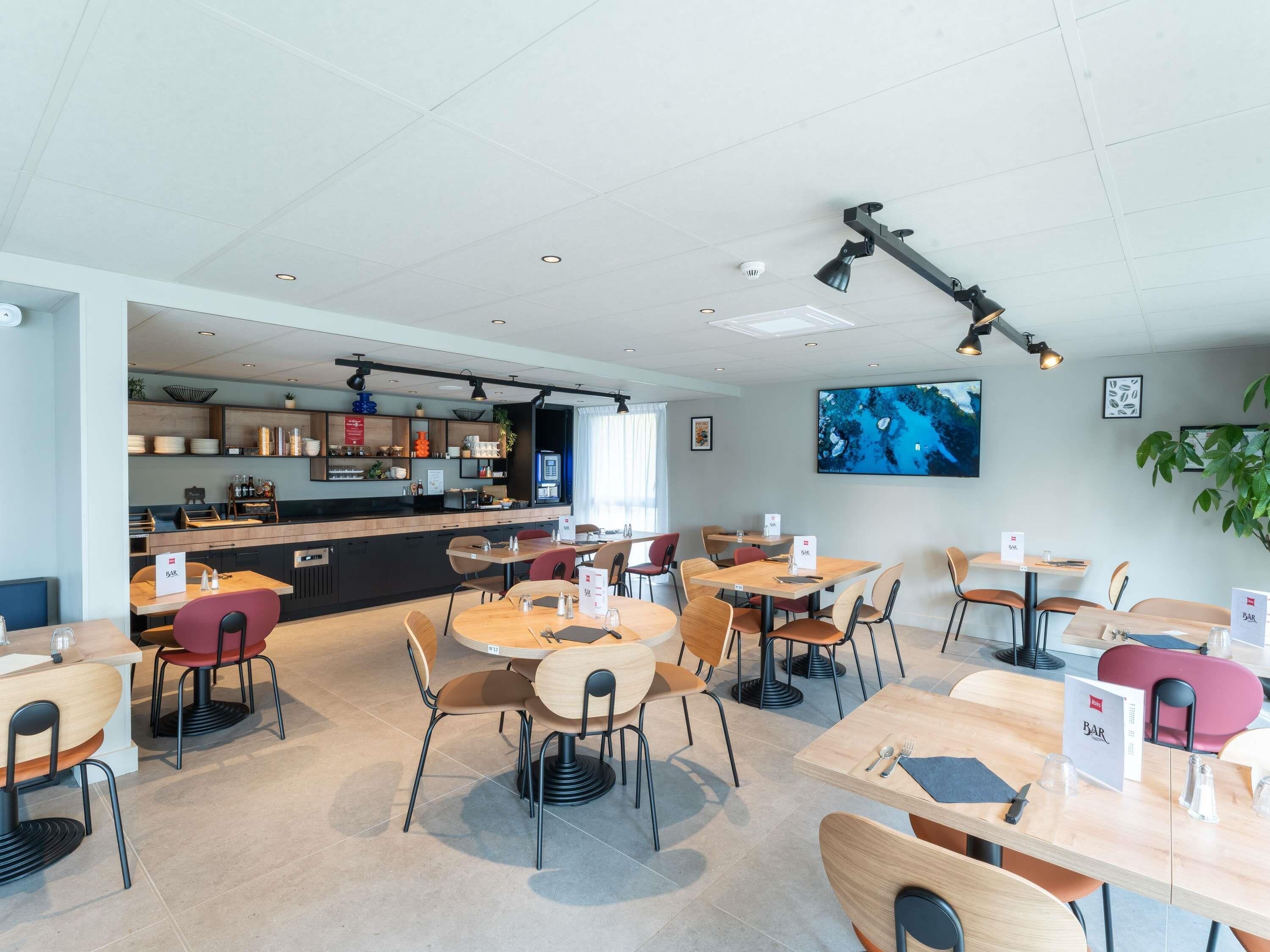A modern restaurant with multiple tables and chairs, a bar area, and a large screen displaying a scenic image.