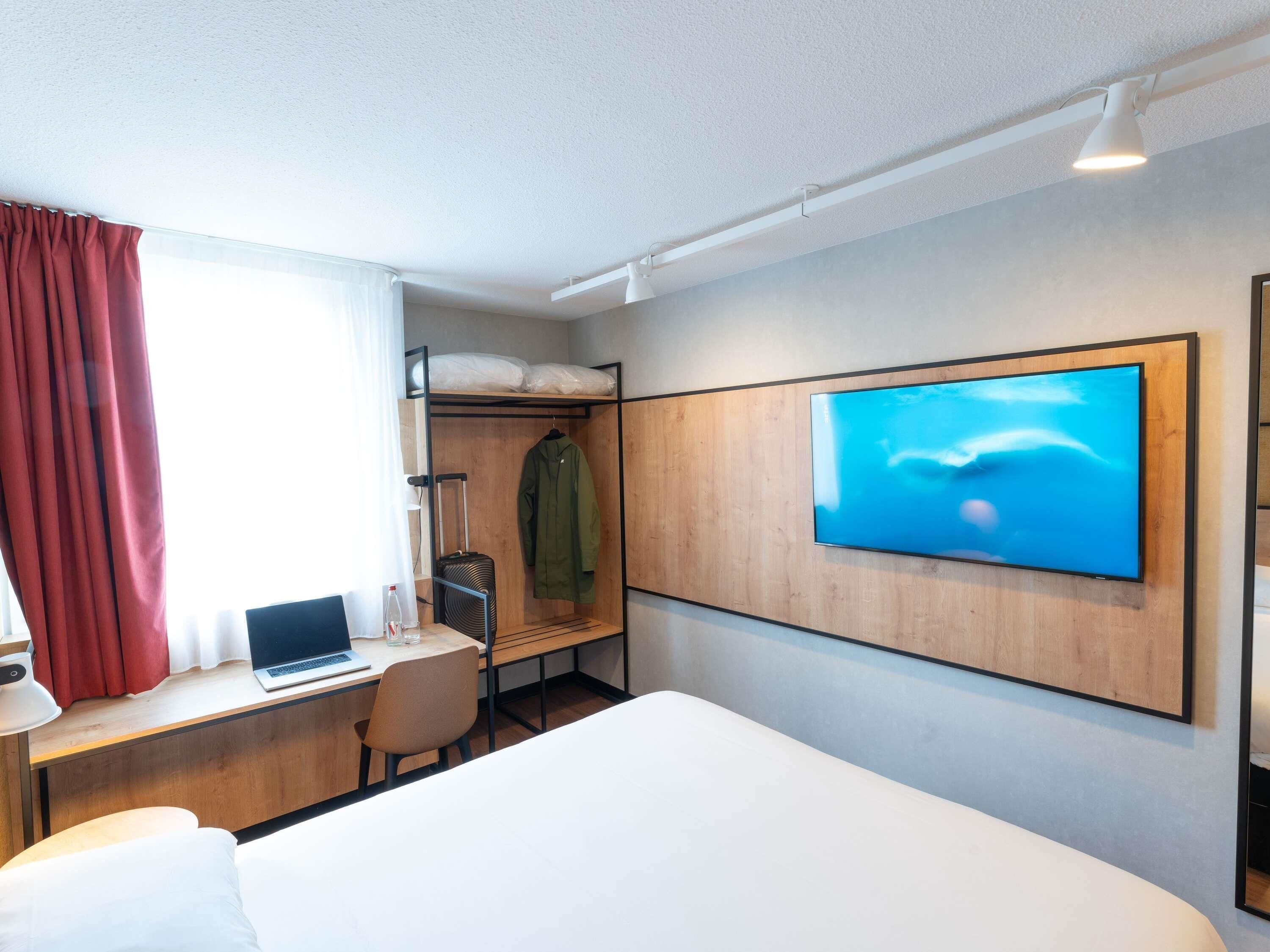 Ett hotellrum med en säng, ett skrivbord med en bärbar dator, en tv som är monterad på väggen och en klädställning med en jacka.