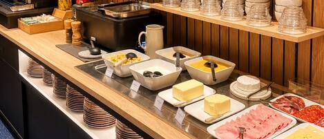 A buffet setup with various food items, condiment containers, and glassware on wooden shelves.