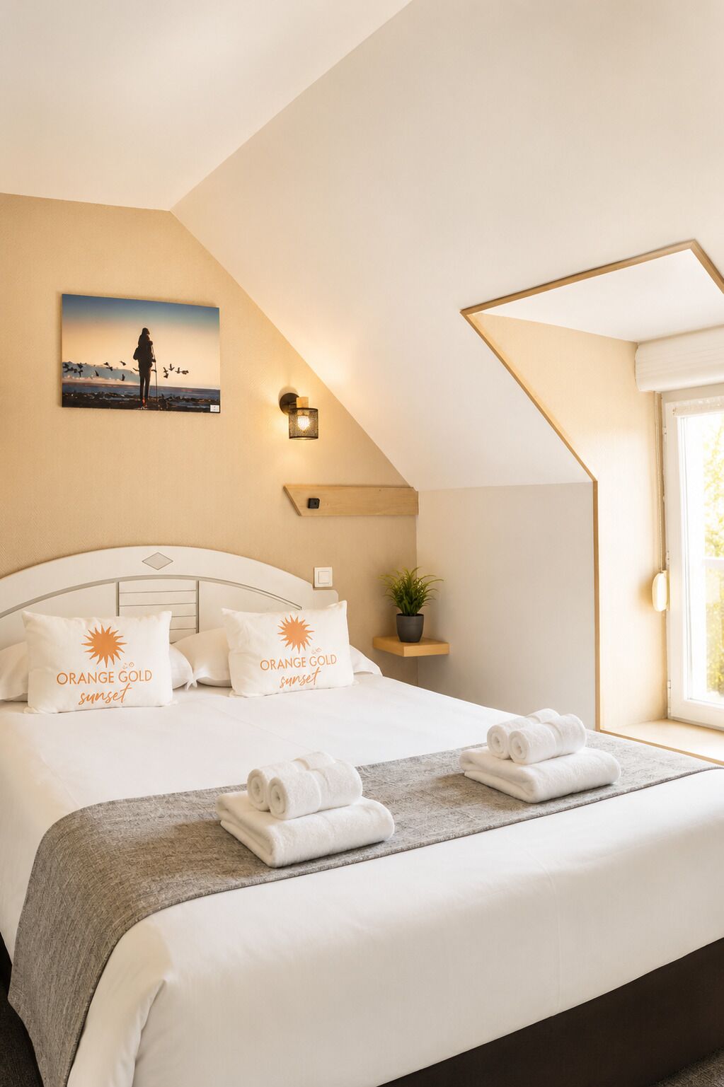 Une chambre d’hôtel moderne avec un grand lit, un tableau représentant une personne avec un oiseau, une lampe fixée au mur, une petite plante et une fenêtre donnant sur un beau paysage.