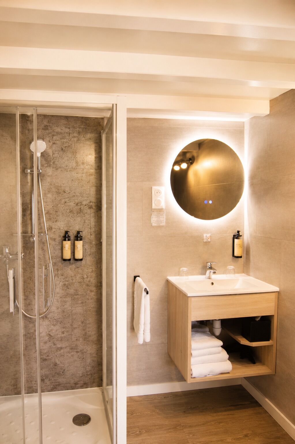 Een moderne badkamer met een douchecabine van glas, een houten wastafelmeubel met een ronde spiegel en een handdoekhouder.
