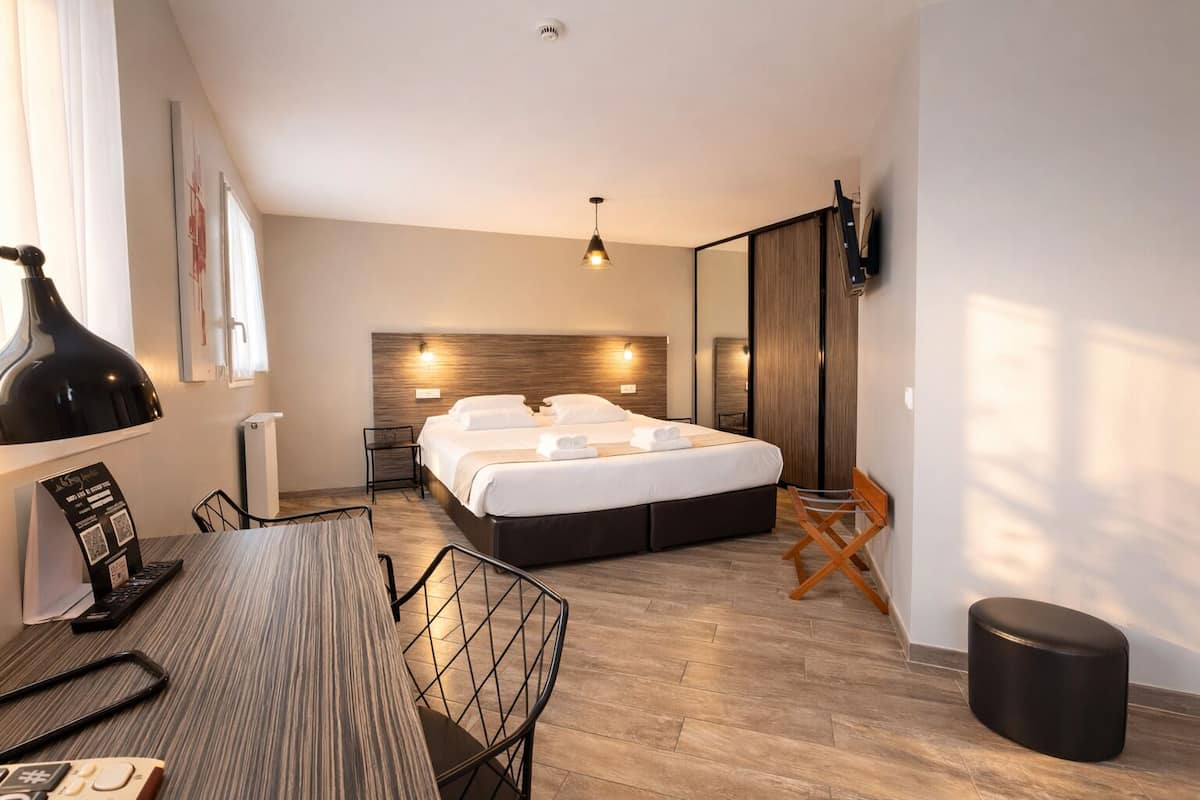 A modern hotel room with a large bed, a desk with a chair, a TV, and a large window.