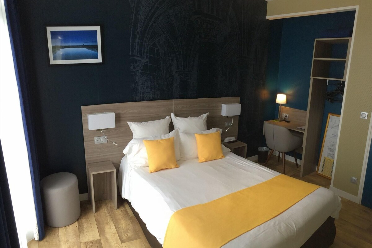 Una habitación de hotel con una cama grande, dos mesitas de noche, un escritorio, una silla, un cesto de basura y una pared con un mural.