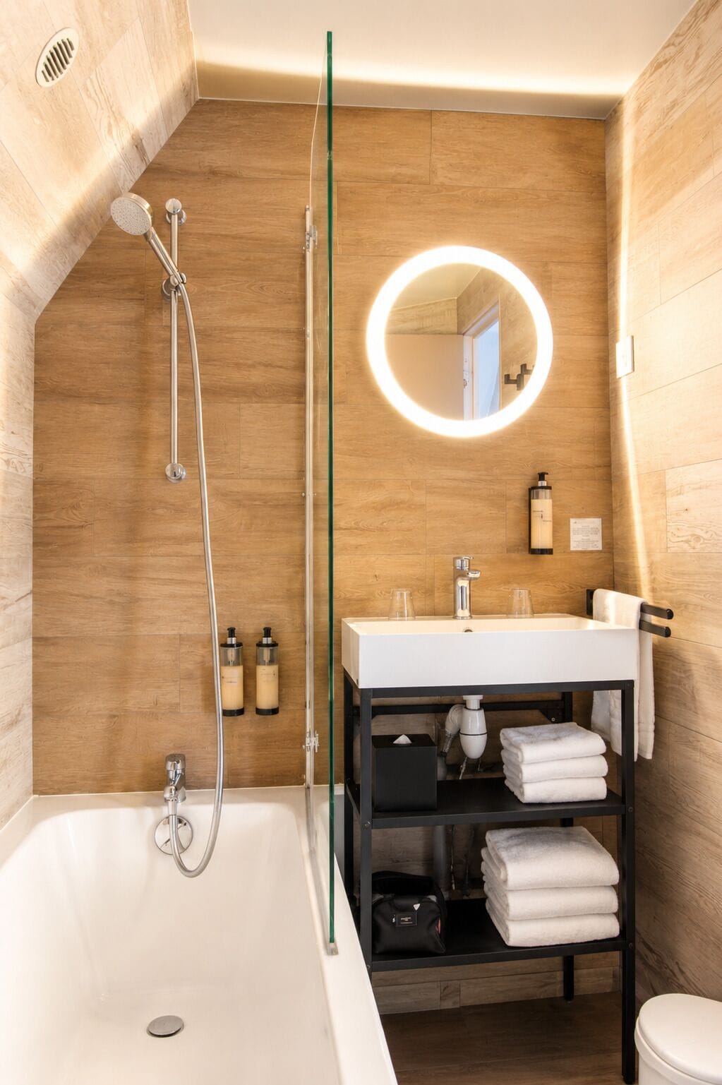 A modern bathroom with a wooden wall, a glass shower enclosure, a white sink, and a mirror with a circular light.