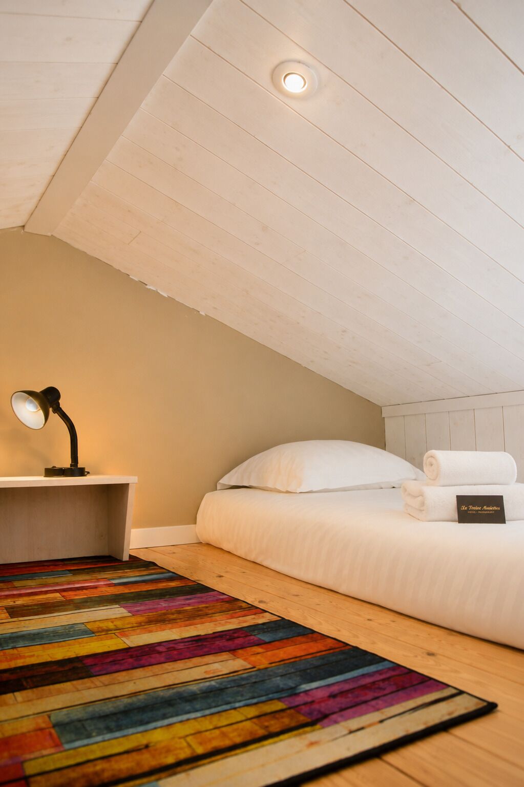 A bedroom with a bed, a desk with a lamp, a colorful rug, and a ceiling light.