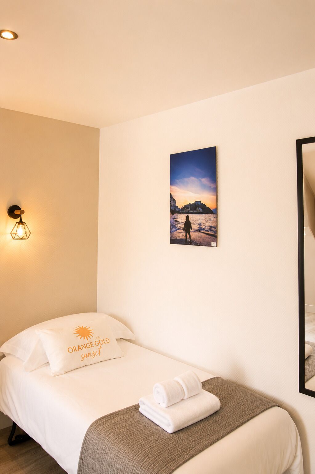 A hotel room with a bed, a wall-mounted picture of a sunset, a wall sconce, and a mirror.
