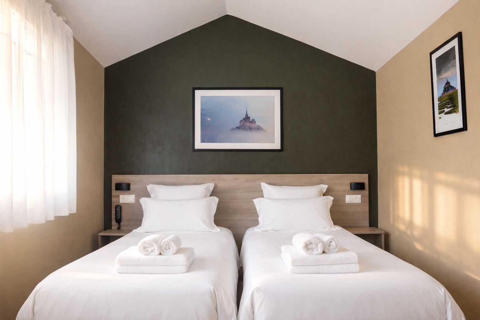Habitación de hotel con dos camas, una pared de acento verde oscuro, un cuadro enmarcado sobre las camas y una ventana con cortinas blancas.