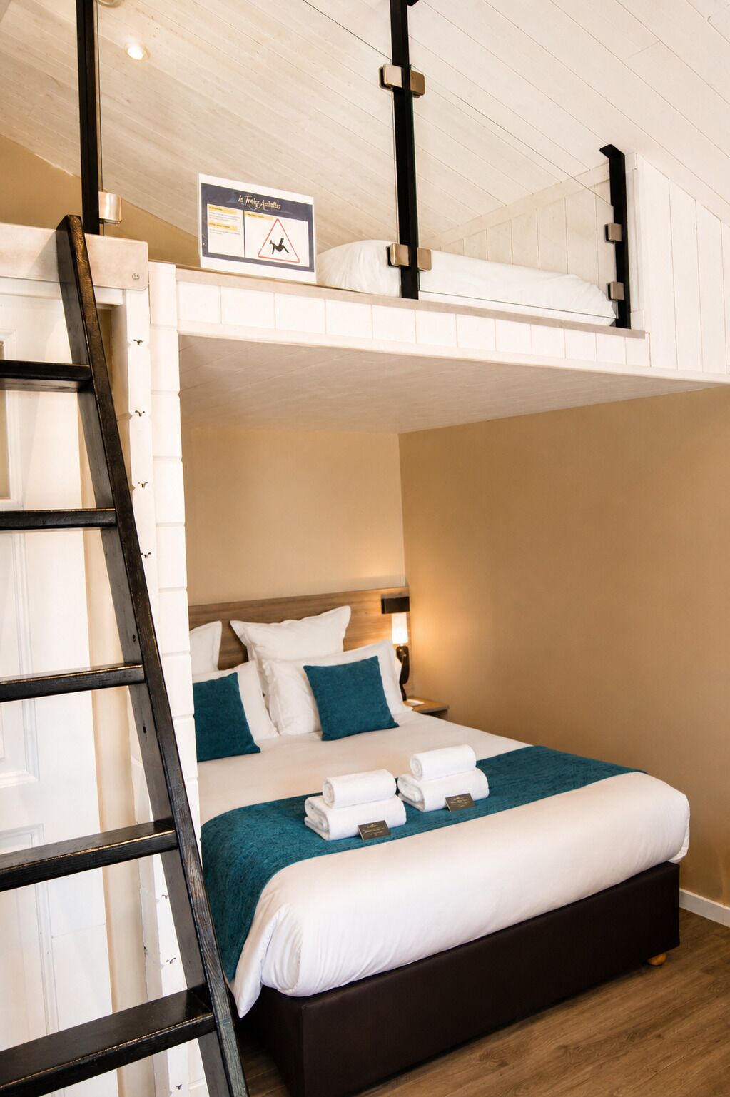 Un lit superposé avec une échelle, une table de chevet avec une lampe et un lit avec du linge de lit blanc et des oreillers turquoise.