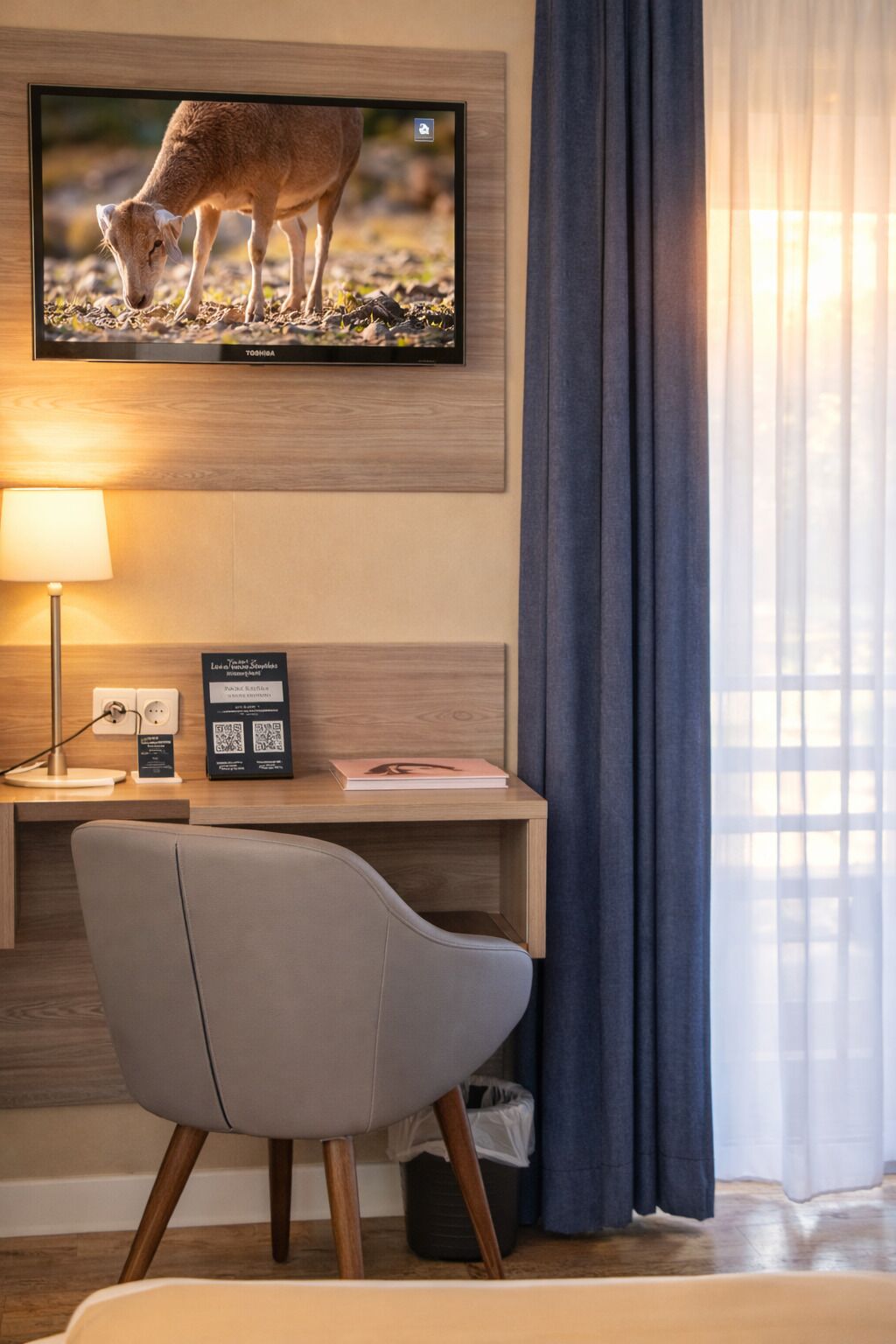 Habitación de hotel con una televisión montada mostrando un ciervo, un escritorio con una lámpara, una silla y un cubo de basura.