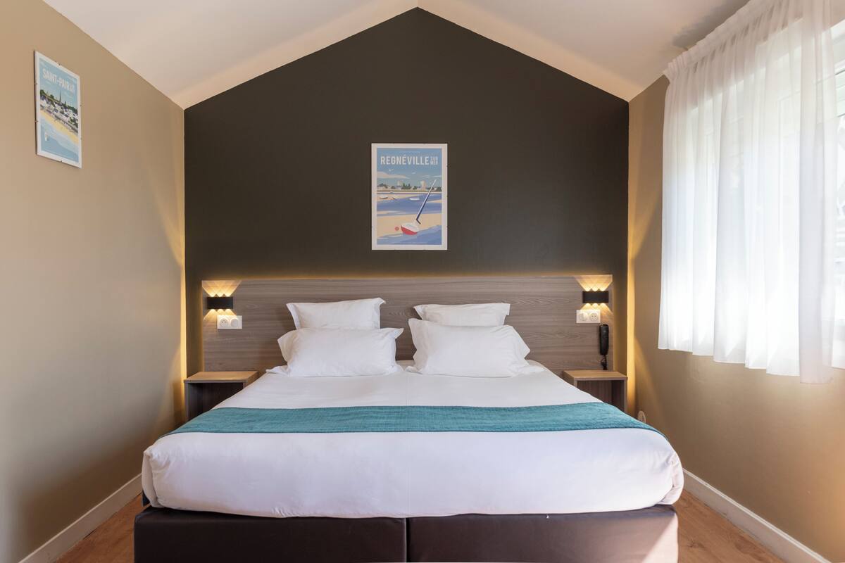 A neatly made bed with white linens and a teal throw, a dark ceiling, and a poster of Regneville.