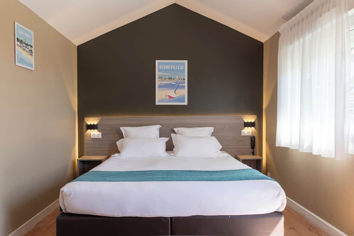 Una cama bien hecha con sábanas blancas y una manta verde aguamarina, un techo oscuro y un cartel de Regneville.