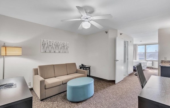 A modern hotel room with a beige sofa, a blue ottoman, a ceiling fan, a painting on the wall, and a view of the city through the window. - Sleep Inn & Suites near Spirit Mountain (Duluth)