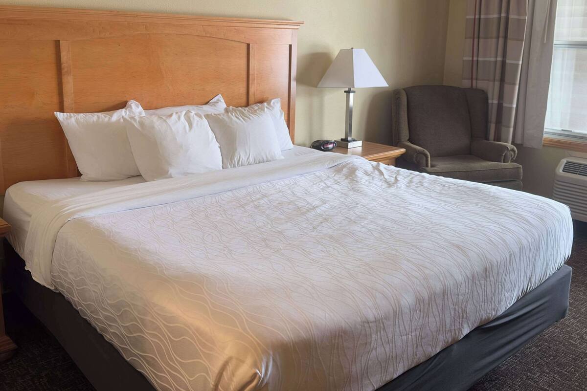 Uma cama bem arrumada com lençóis brancos e cabeceira de madeira, uma luminária de cabeceira e uma cadeira em um quarto de hotel.