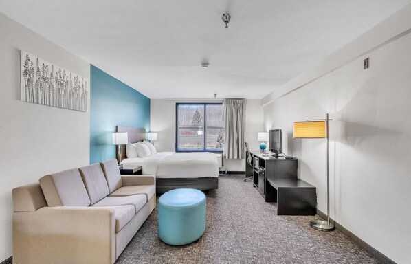 Habitación de hotel con cama, sofá, escritorio, lámpara y ventana con vista a árboles.