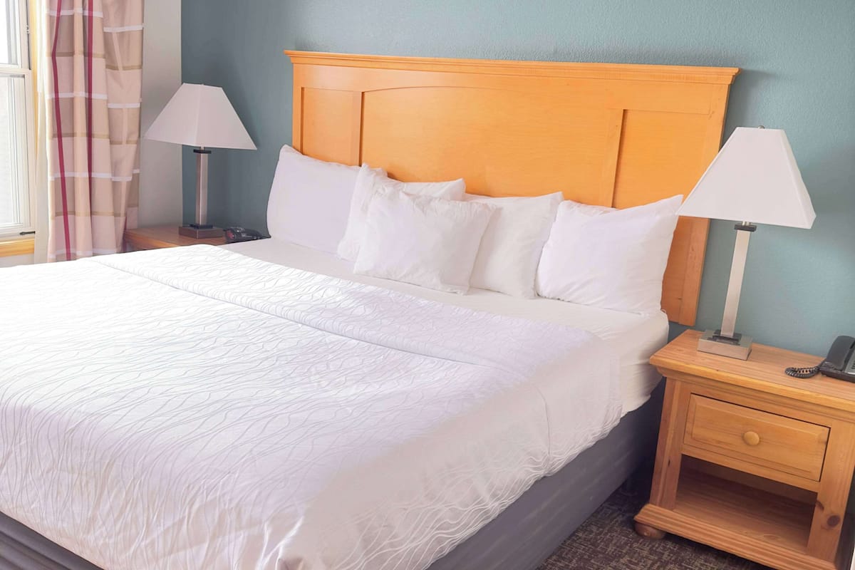 Cama bem arrumada com lençóis brancos, cabeceira de madeira e duas mesas de cabeceira com abajures.