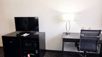 A hotel room with a flat-screen TV, a desk with a lamp, a black office chair, and a minibar.