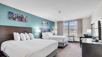 Quarto de hotel com duas camas, uma escrivaninha com computador, uma janela com cortinas e obras de arte nas paredes.